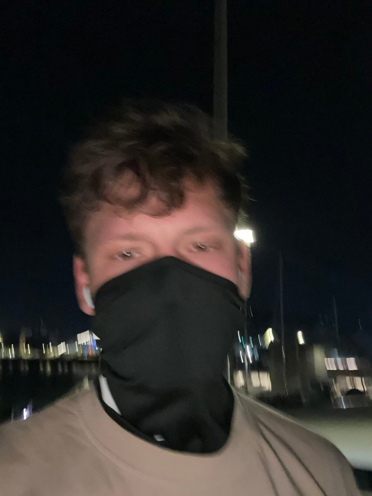 Blurry nighttime selfie of a man wearing a black mask and AirPods, with city lights and a dark sky in the background.