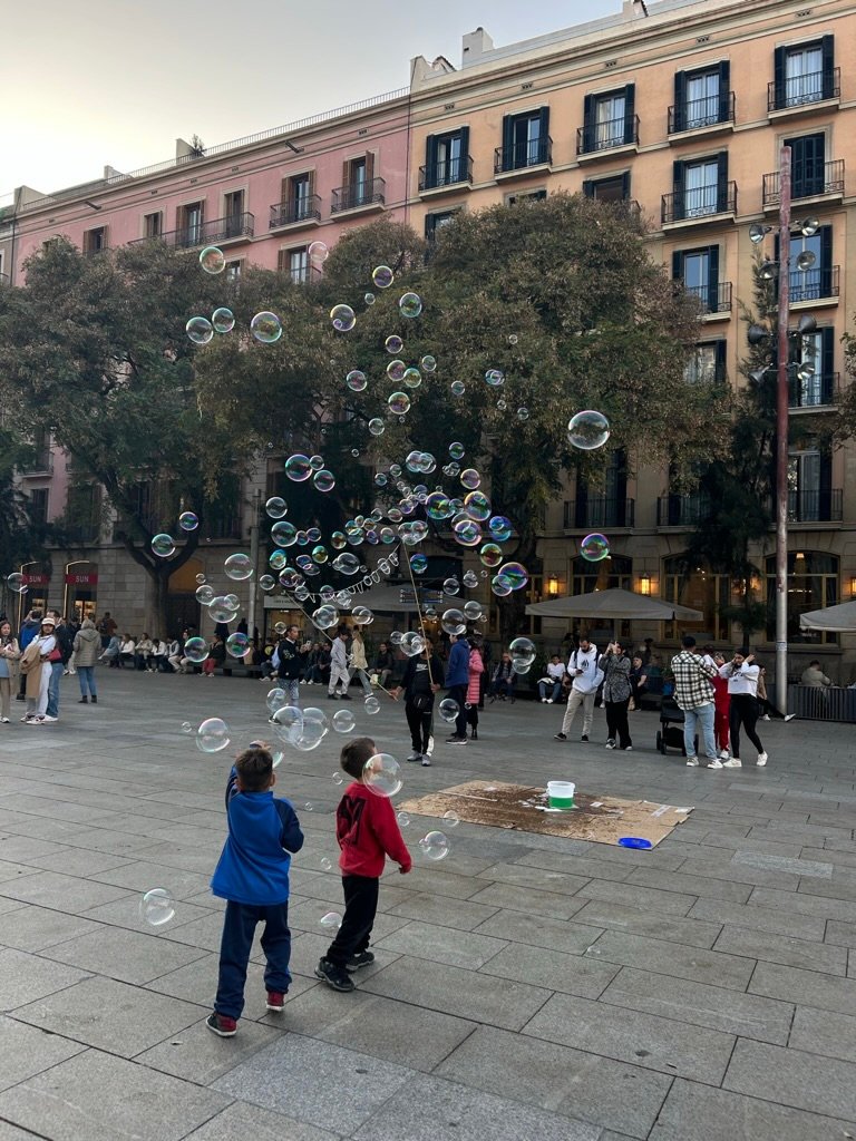 Children play with floating soap bubbles in a lively city square, surrounded by people and colorful buildings at dusk.