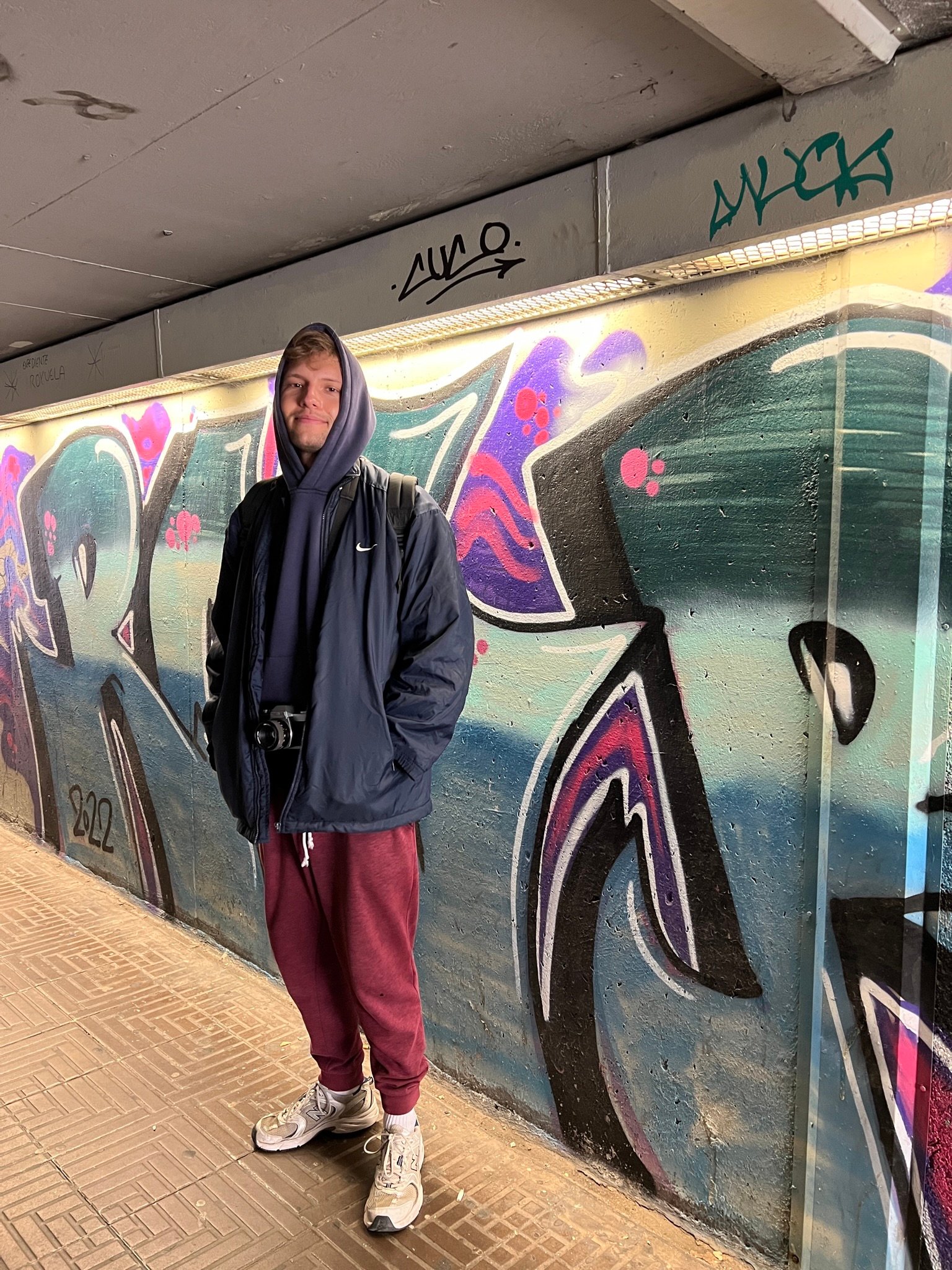 Young man in a hoodie and jacket standing in front of a brightly colored graffiti wall in an urban underpass.