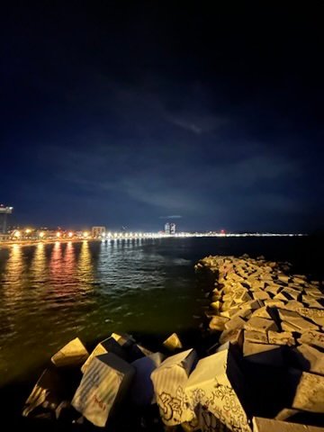Nighttime view of a rocky jetty leading into the ocean, with city lights glowing across the shoreline.