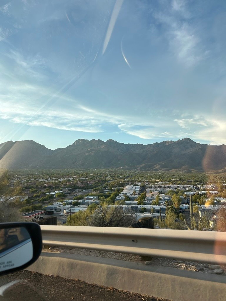 A view from a car window showing a southwestern town nestled at the foot of rugged mountains under a soft blue sky.