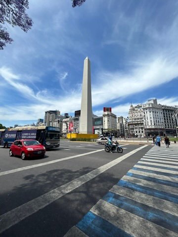 A tall obelisk monument rises in the center of a busy city intersection with cars, buses, and people crossing beneath a vibrant blue sky