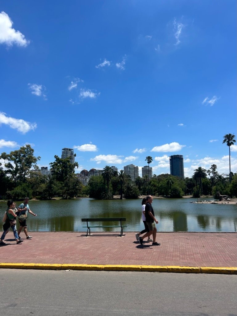 A sunny urban park with a lake, palm trees, and city buildings in the background, as people walk along a pathway near an empty bench