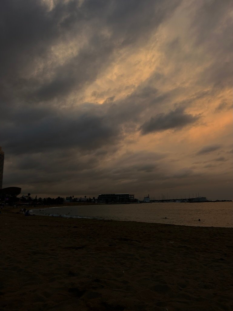 A moody beach scene under dramatic, dark clouds at sunset, with distant silhouettes of buildings and swimmers in the water.