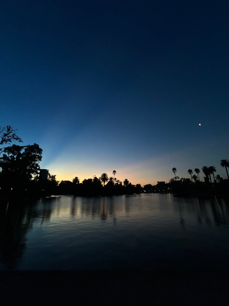 A peaceful lake at dusk with dark silhouettes of palm trees against a deepening blue and orange sky, with a small crescent moon visible