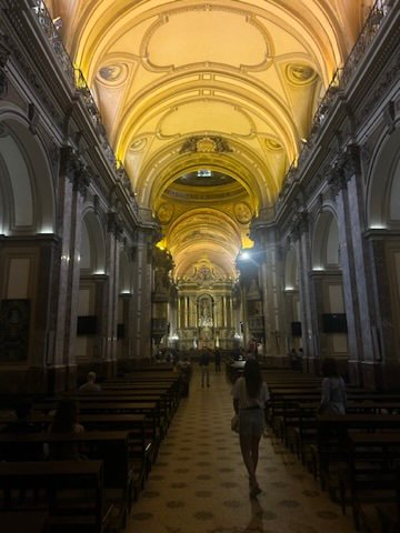 The interior of a beautifully lit cathedral with golden arched ceilings, marble columns, and rows of wooden pews leading to an ornate altar