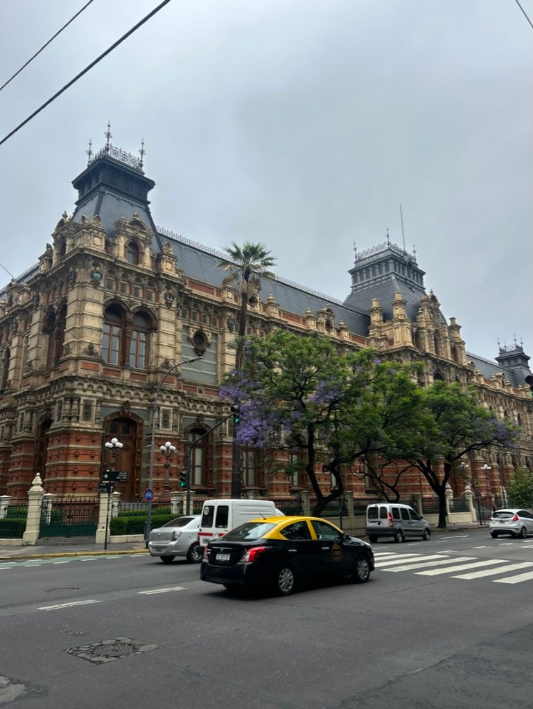 A historic, ornate building with detailed architecture and towers under a cloudy sky, with cars and taxis passing by on the street in front