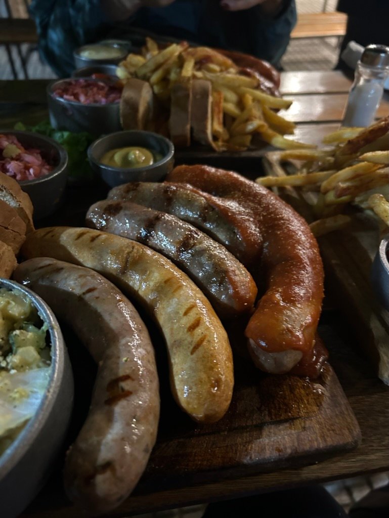 A wooden platter filled with grilled sausages, French fries, and dipping sauces on a table at an outdoor restaurant.