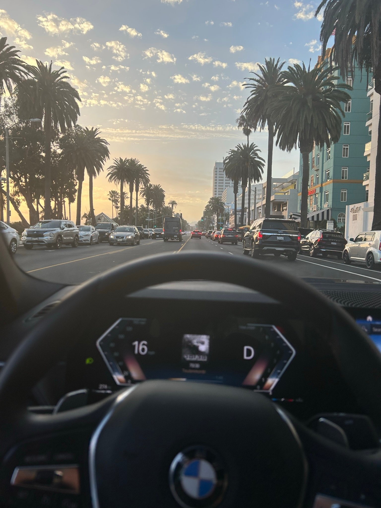 View from inside a BMW looking out at a palm tree-lined street at sunset with light traffic.