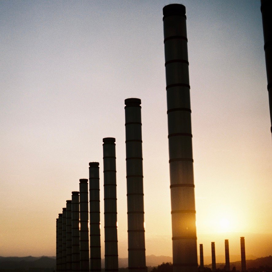 A series of tall industrial smokestacks silhouetted against a golden sunset sky, lined up in a row fading into the distance