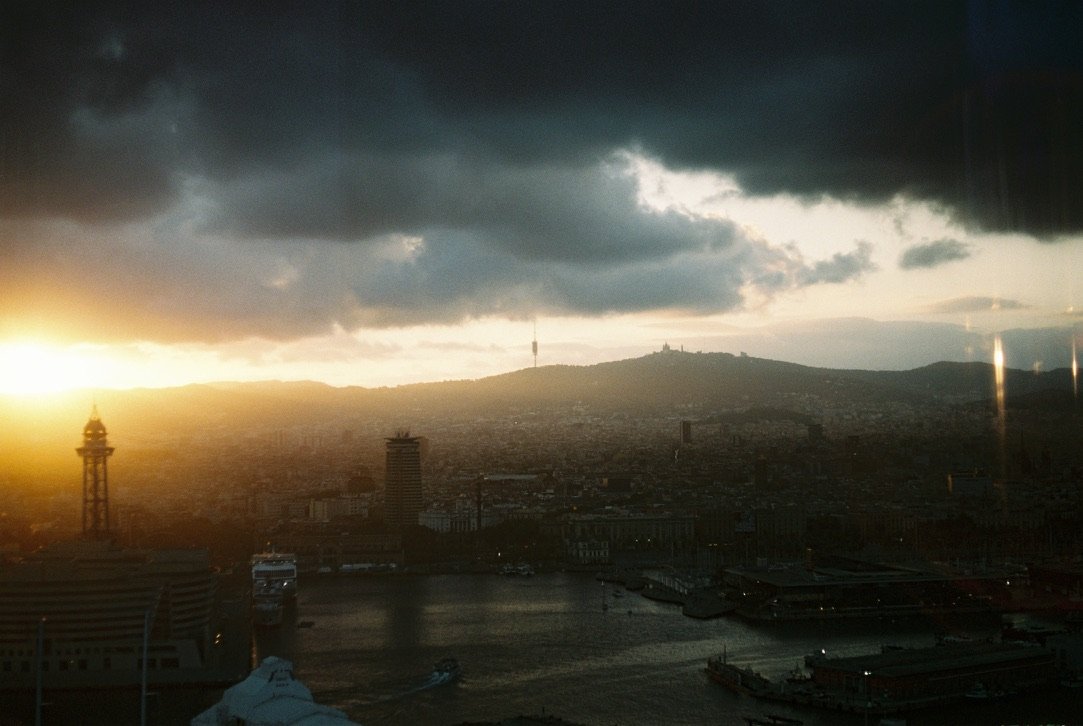 A cityscape at sunset with dramatic dark clouds over mountains and a harbor, as sunlight breaks through and reflects off the water