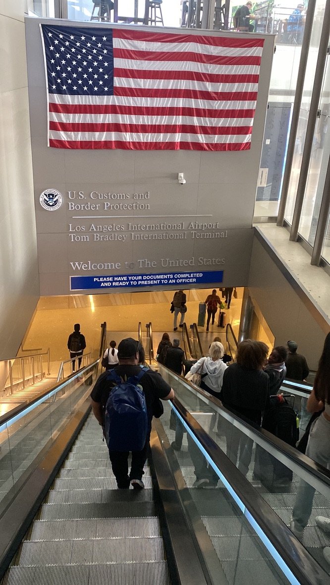 Travelers descend an escalator at LAX under a large American flag and U.S. Customs and Border Protection sign