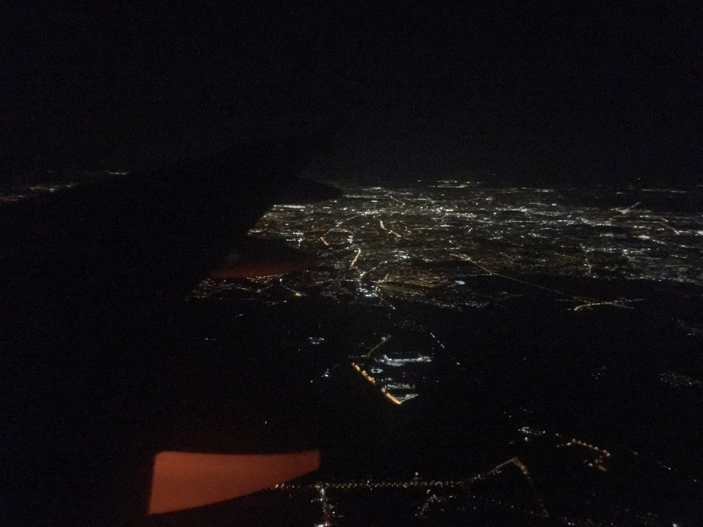 A nighttime view from an airplane window showing the glowing city lights below, with the airplane wing faintly visible against the dark sky