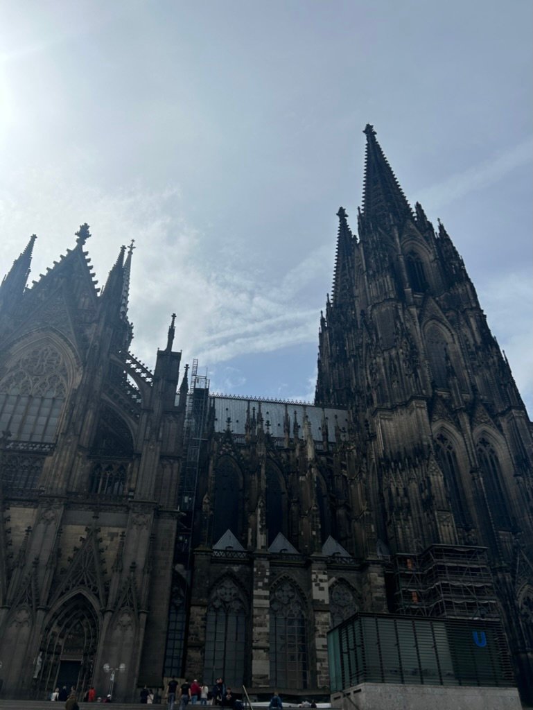A towering Gothic cathedral with dark spires reaching into a cloudy sky, with people walking near its base, highlighting its grand and intricate architecture