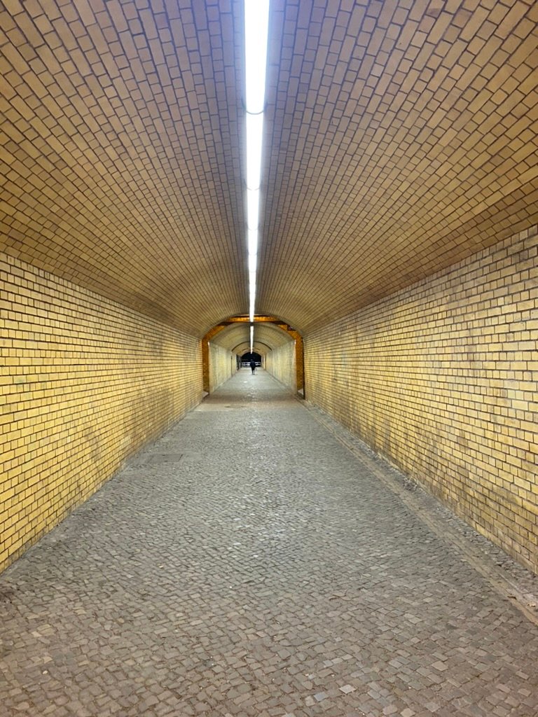 A long, yellow brick tunnel with a cobblestone floor, illuminated by a single bright fluorescent light running along the ceiling