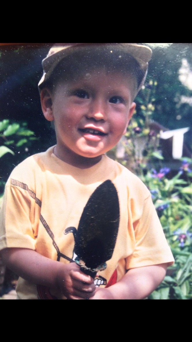 A smiling young boy holding a garden trowel, wearing a yellow shirt and cap, standing outdoors with plants in the background
