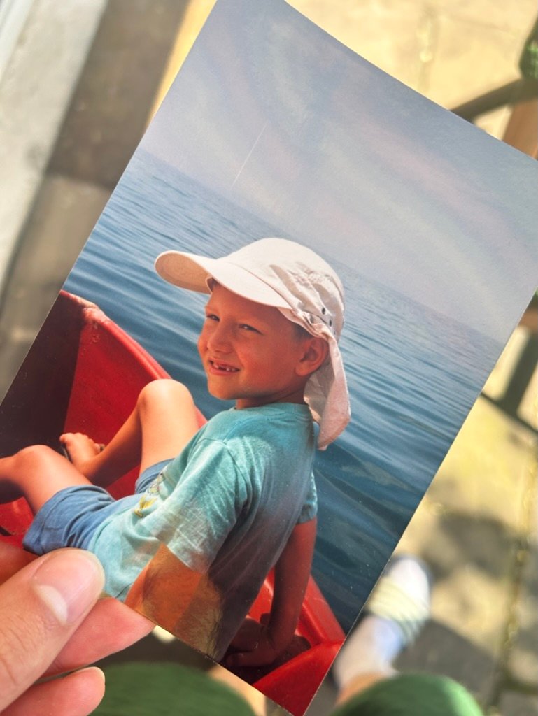 A printed photo of a smiling young boy wearing a light hat and blue shirt, sitting in a red boat on calm blue water, held by a hand