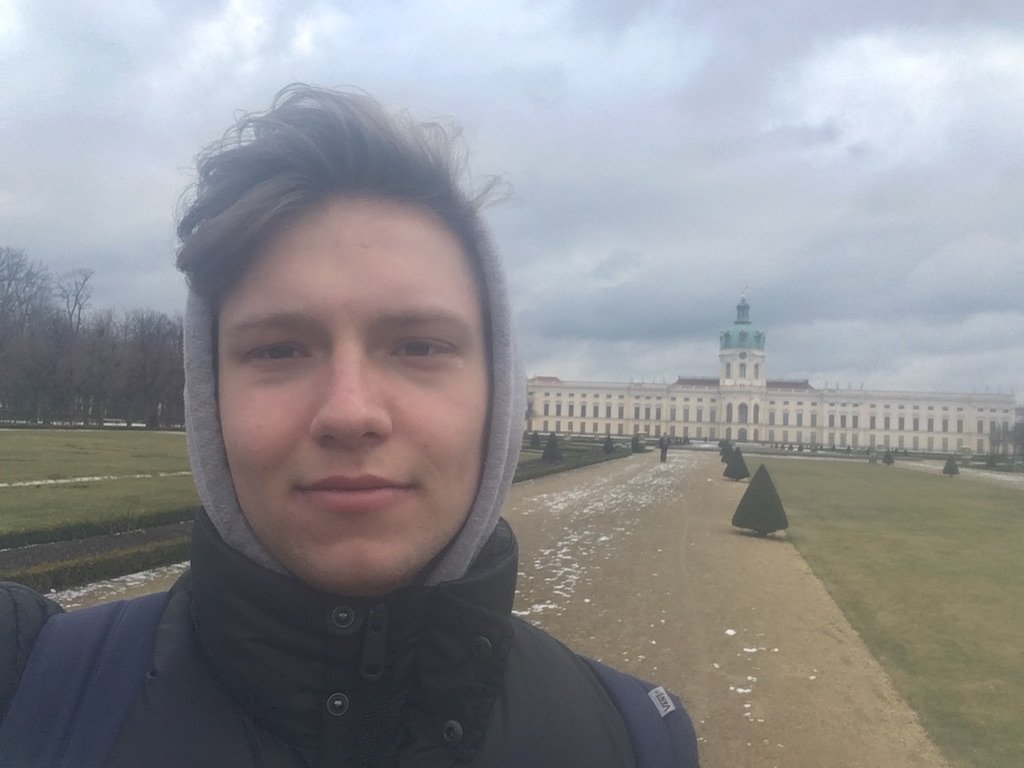 Young man wearing a hooded jacket taking a selfie in front of Charlottenburg Palace on a cold, cloudy day