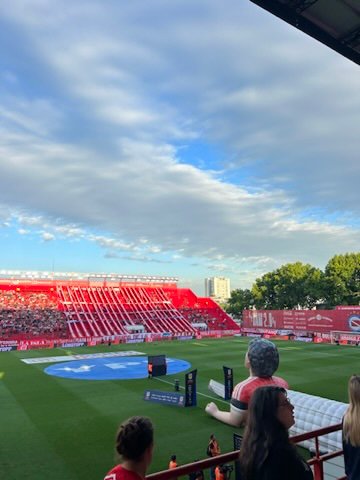 A vibrant soccer stadium with red stands filled with fans, a partly cloudy sky above, and people watching from the stands near the field
