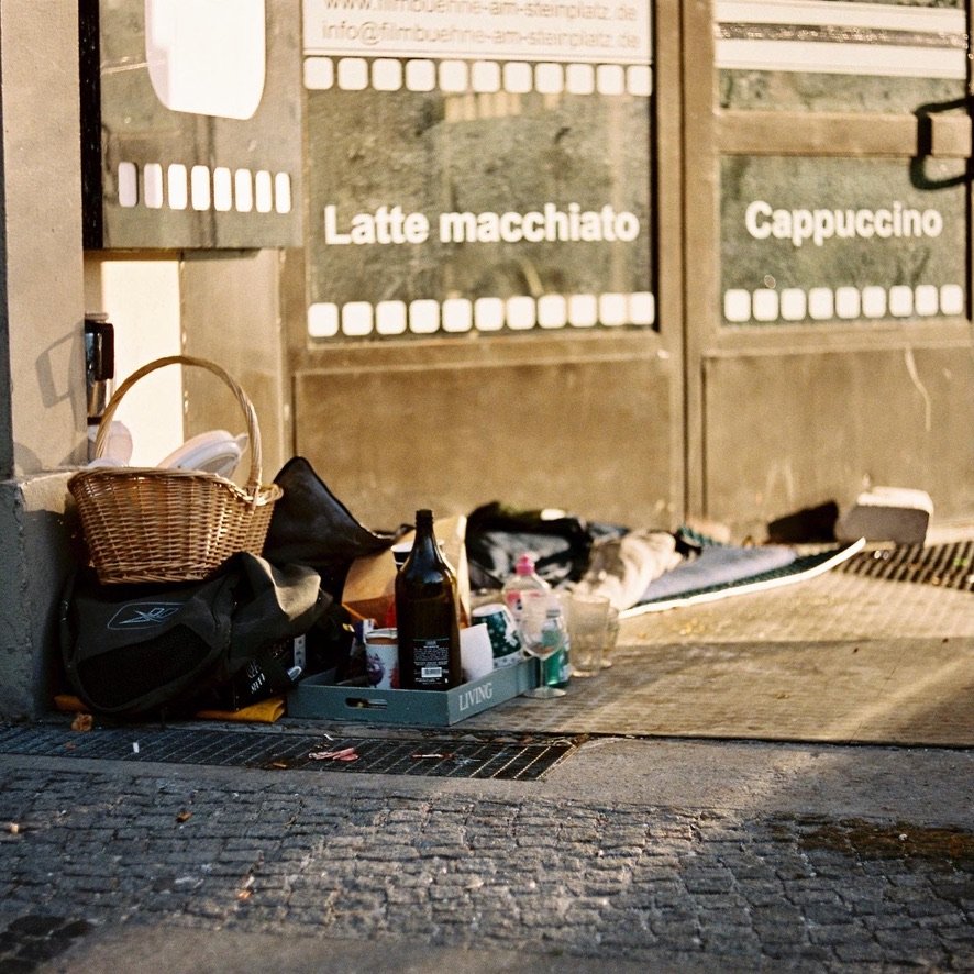 A makeshift setup of belongings including a basket, bottles, and bedding outside a cafe with signs for "Latte macchiato" and "Cappuccino"