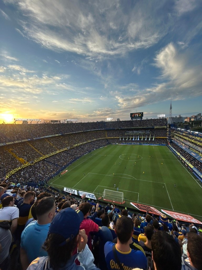 A packed soccer stadium at sunset with a clear view of the field, players, and a vibrant sky above, as fans watch the match with excitement