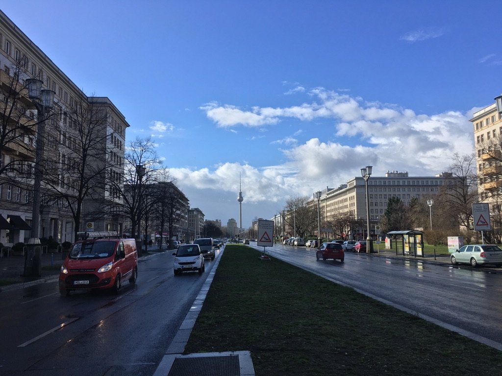A city street in Berlin with wet pavement, cars, tall buildings, and the Fernsehturm (TV Tower) visible in the distance under a blue sky with scattered clouds