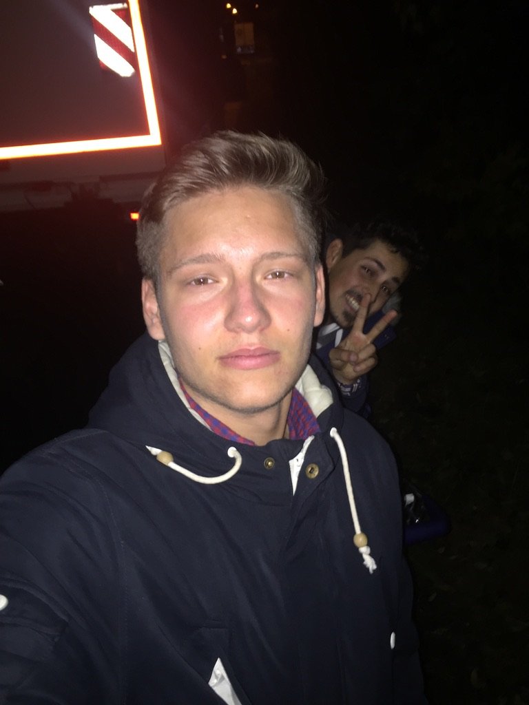 Two young men taking a nighttime selfie outdoors, one in front wearing a hooded jacket, the other in the background flashing a peace sign