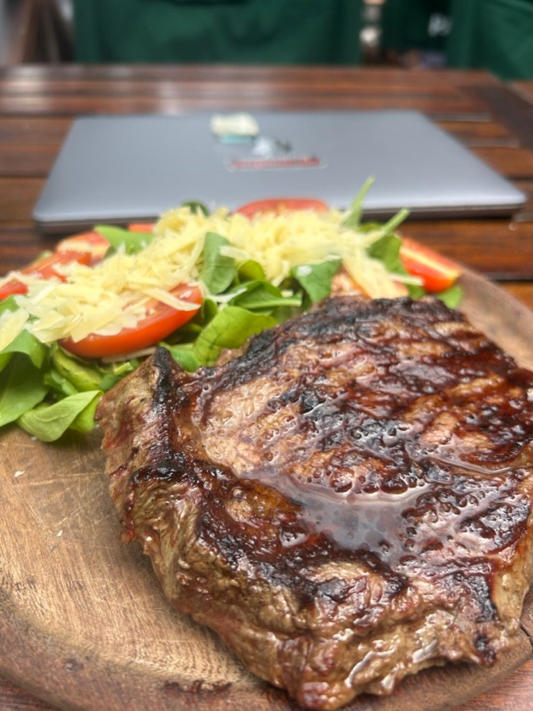 A grilled steak served with fresh salad on a wooden plate, with a laptop blurred in the background