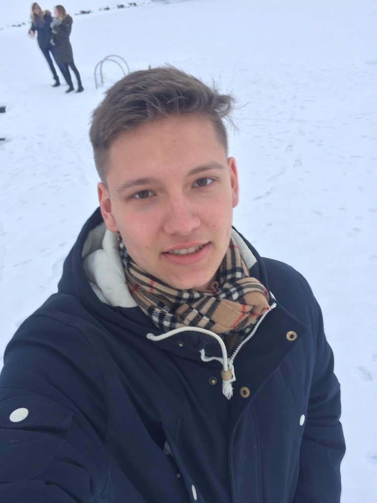 A young man in a dark winter coat and plaid scarf smiles in a snowy landscape with two women in the background