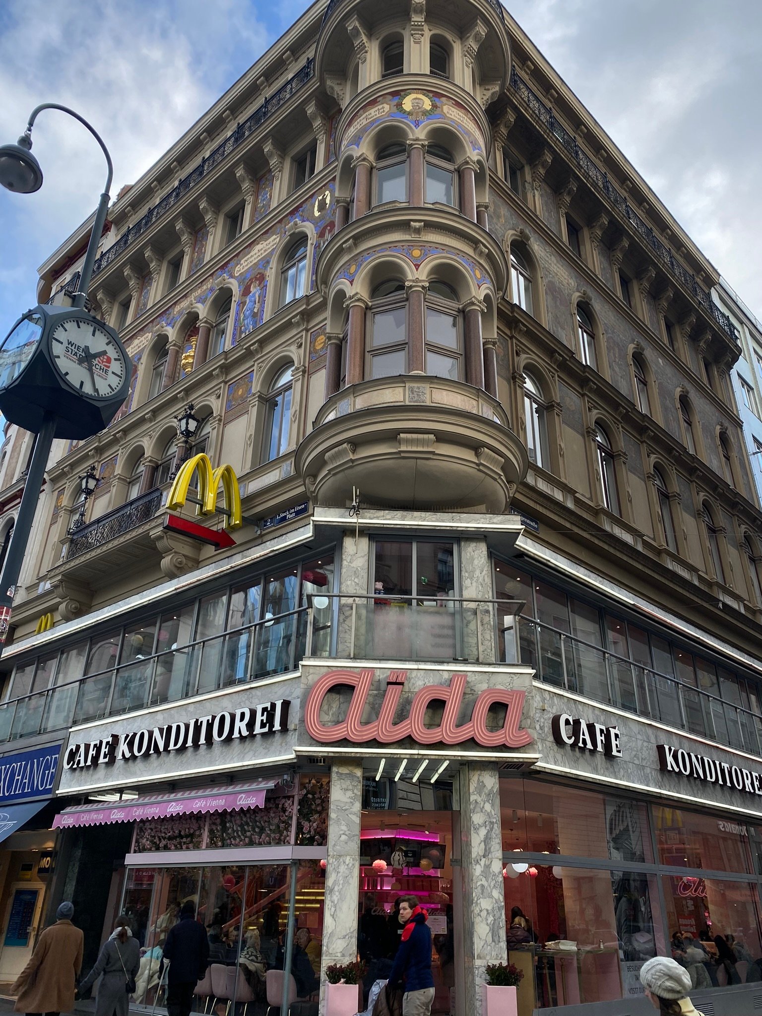 A richly decorated European building with a corner café named Aida, ornate balconies, and religious-style murals above a McDonald's sign