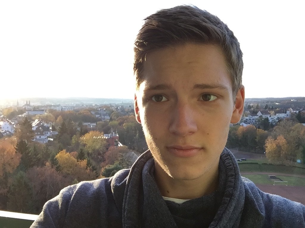 A young man taking a selfie on a balcony during golden hour, with a scenic town and autumn trees in the background
