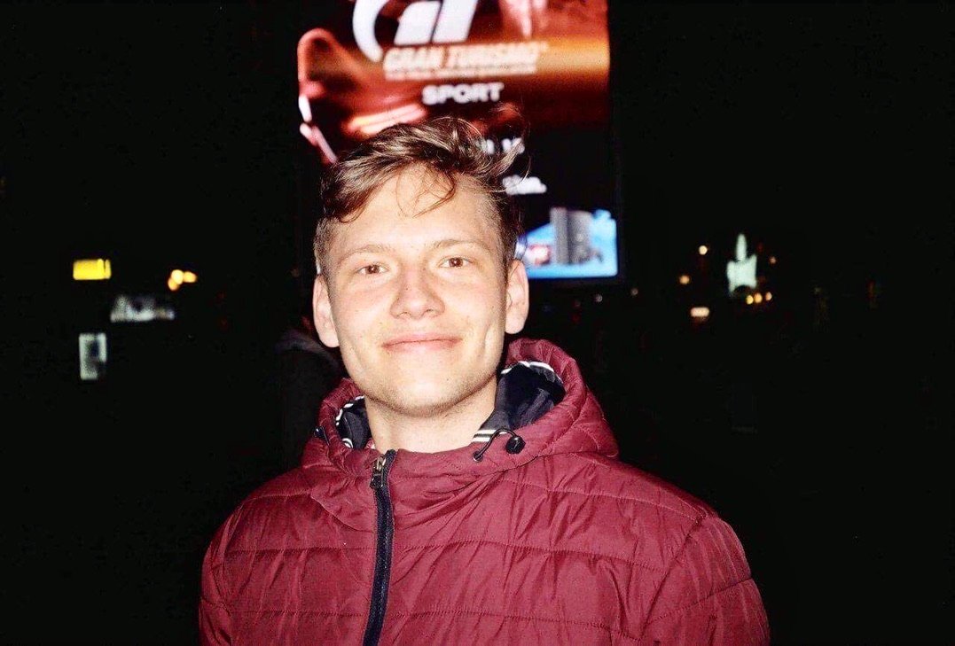 A young man in a maroon jacket smiling at night with a glowing billboard behind him