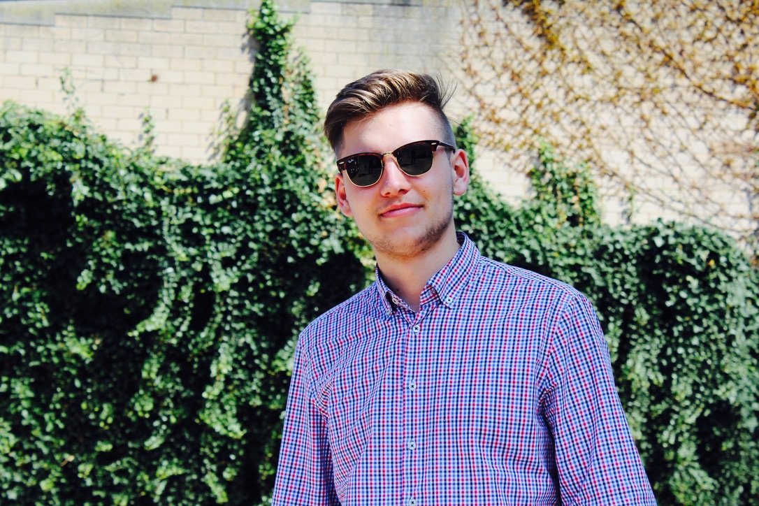 Young man in sunglasses and a checkered shirt standing outdoors in front of a sunlit ivy-covered wall