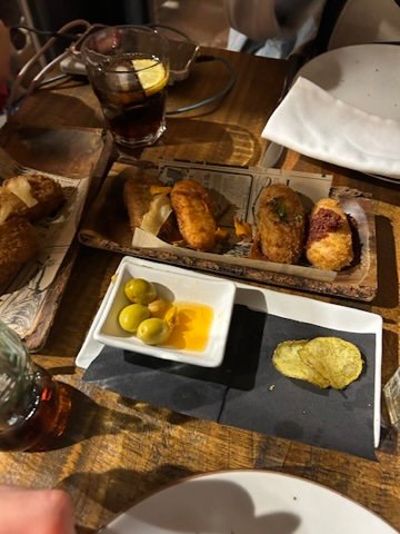 A wooden table with assorted tapas including croquettes, olives in oil, potato chips, and drinks in a cozy dining setting