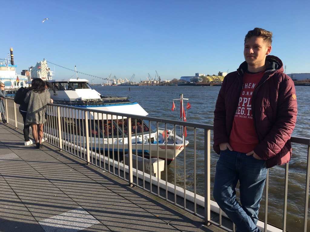 Young man in a red jacket standing by a harbor railing on a sunny day with boats and industrial cranes in the background