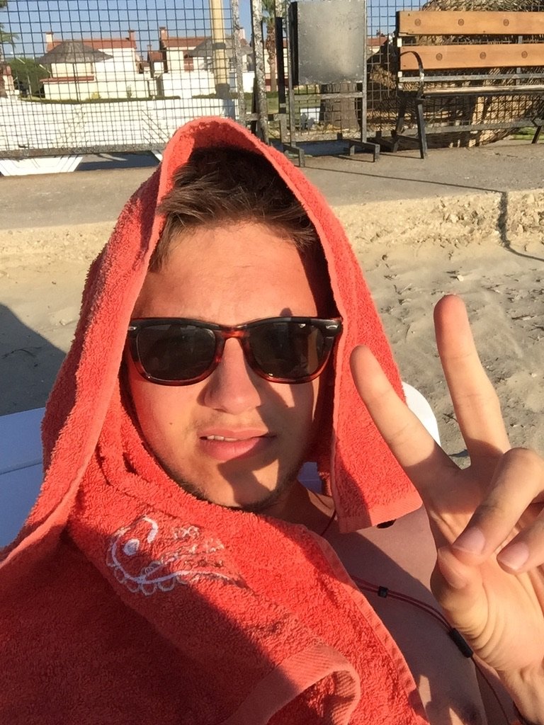 A young man wearing sunglasses and a red towel on his head makes a peace sign while relaxing in the sun.