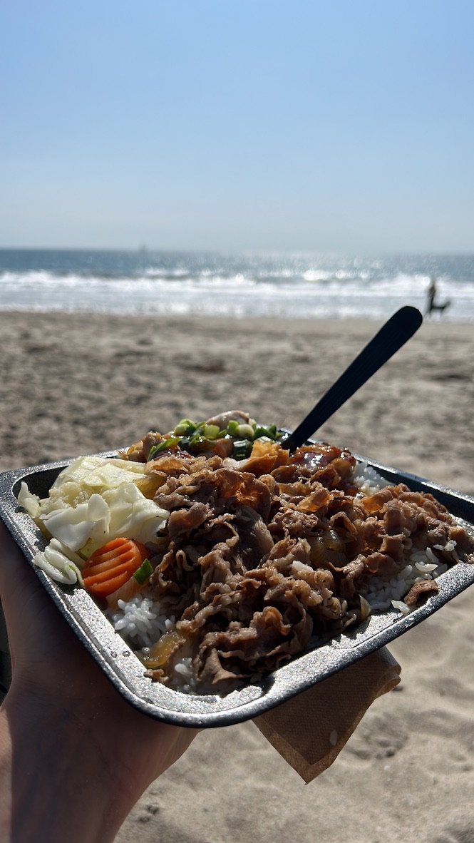A hand holds a tray of rice, vegetables, and meat in focus, with a sandy beach and sparkling ocean waves in the background.