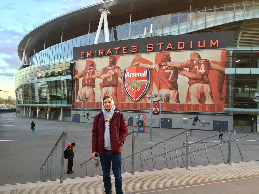 A young man stands in front of Emirates Stadium, home of Arsenal Football Club, with a mural of legendary players behind him.