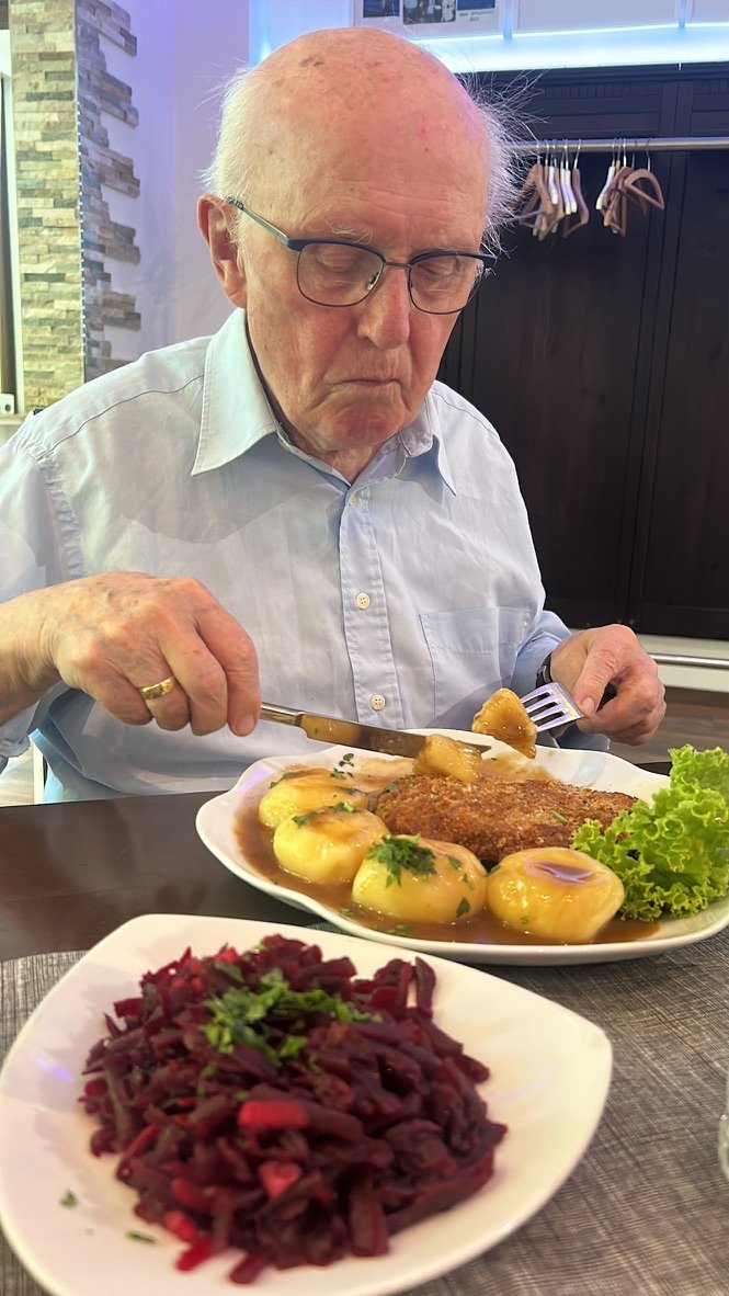 An elderly man in glasses and a light blue shirt enjoys a hearty meal of gravy-covered potatoes, schnitzel, and a side of beet salad.