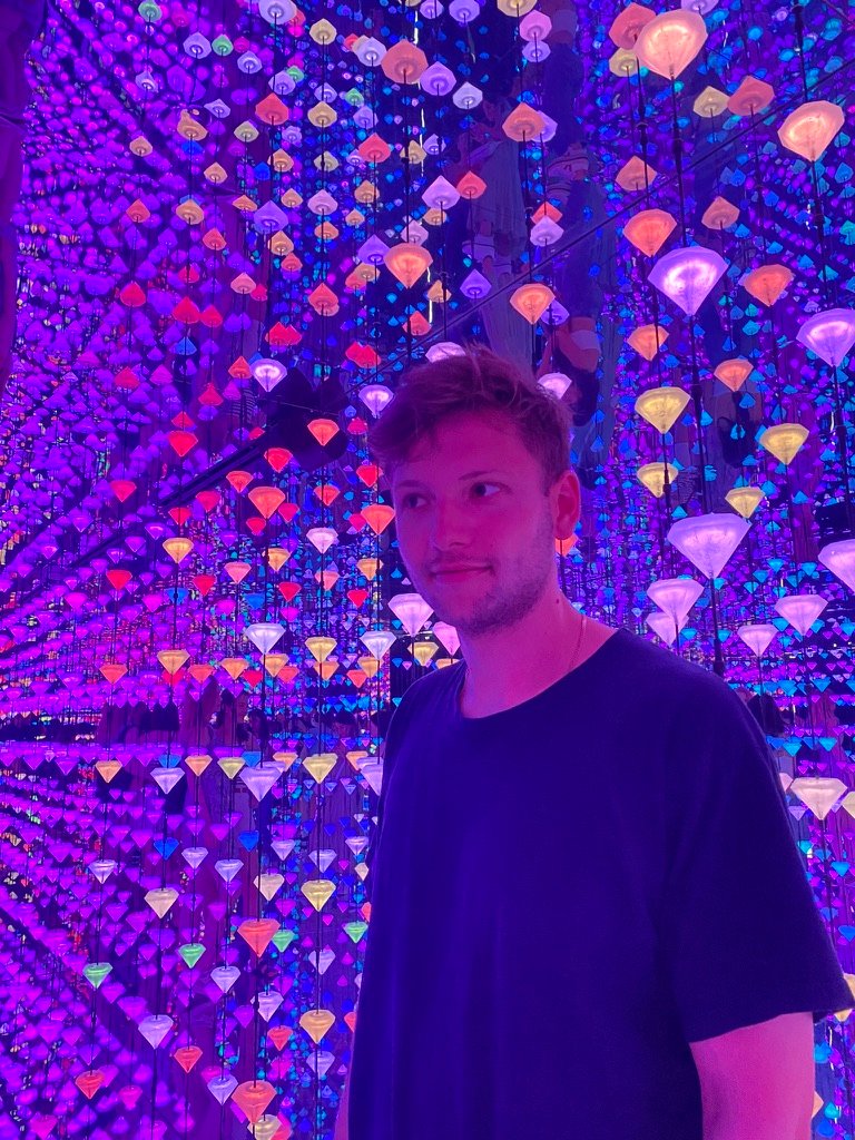 A young man stands in a room filled with hanging, glowing, multicolored diamond-shaped lights, creating a surreal, dreamlike atmosphere.