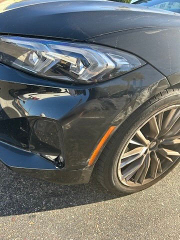 A close-up of a black car’s front bumper and wheel, marked by scrapes and dents.