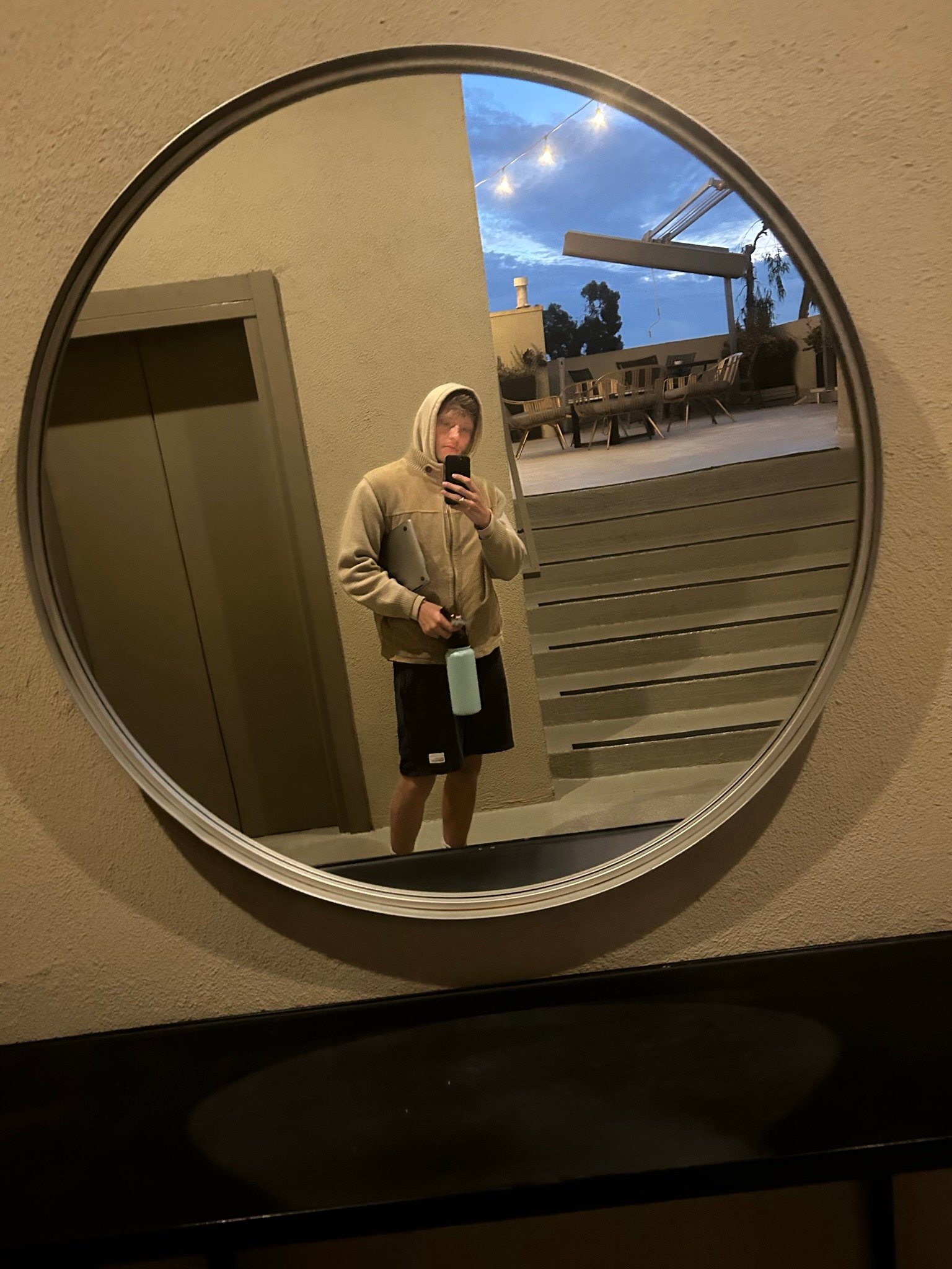 A man in a hoodie takes a mirror selfie at dusk, holding a water bottle, with outdoor lights glowing behind him.