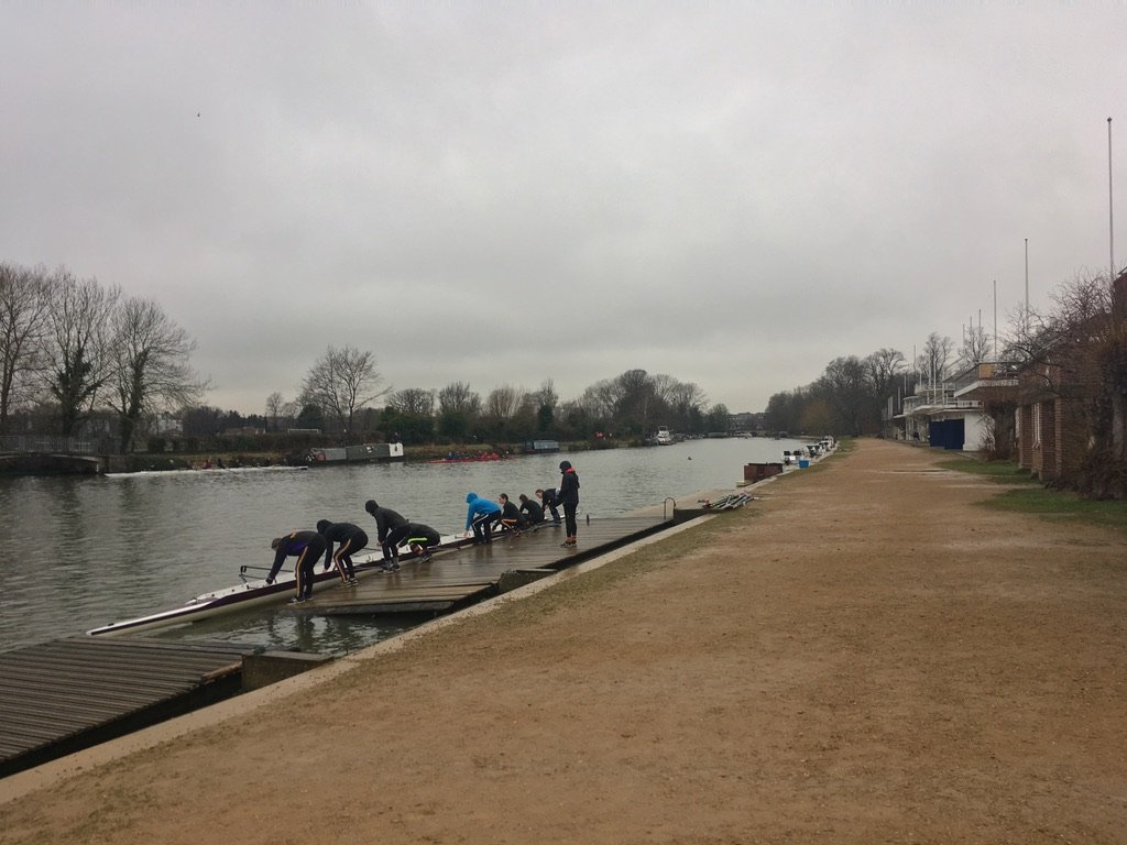 A rowing team prepares their boat on a cloudy day by the riverside dock