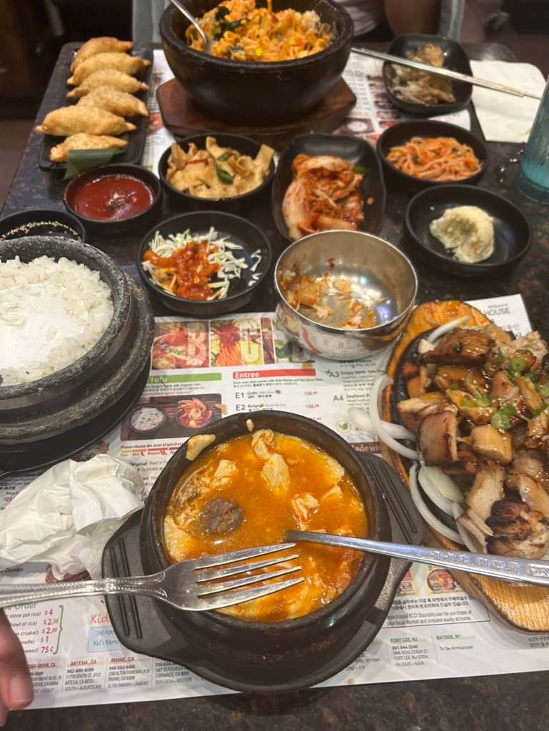 A vibrant Korean meal spread across a table, including kimchi, rice, soup, dumplings, and grilled meats.
