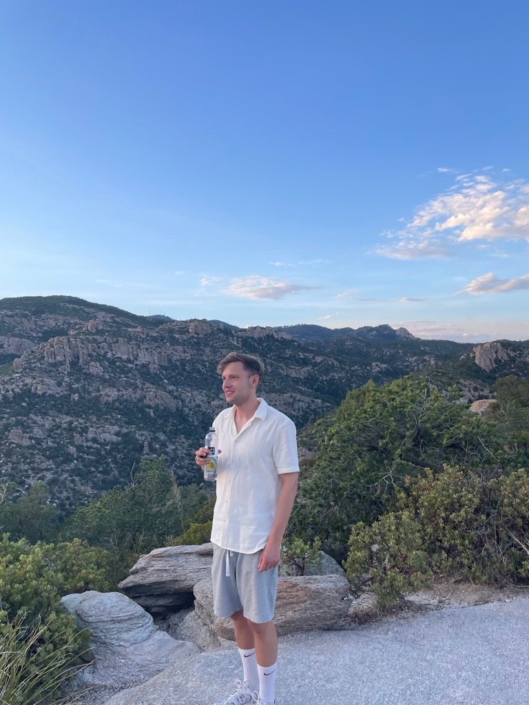 A young man stands on a rocky overlook, holding a bottle, surrounded by mountains and trees under a bright blue sky.