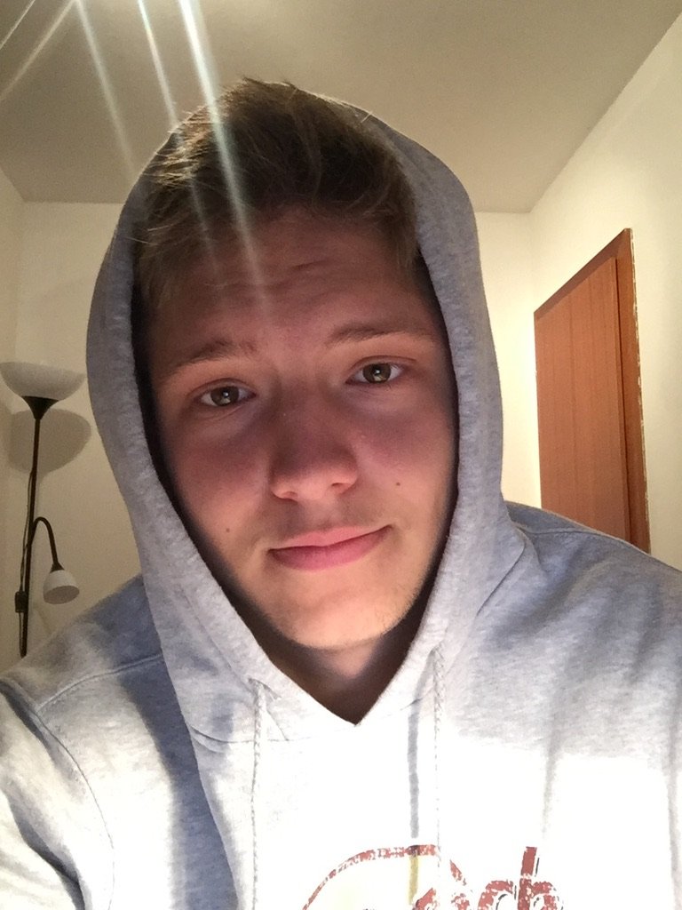 A young man wearing a gray hoodie sits indoors under warm lighting, looking calmly into the camera