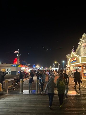 eople walking along a brightly lit pier at night, with shops and lights shining against the dark sky.