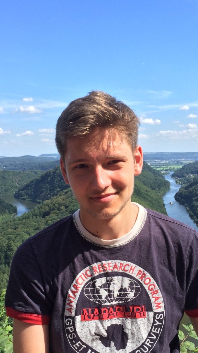 A young man stands smiling in front of a sweeping river bend surrounded by green hills under a bright blue sky.