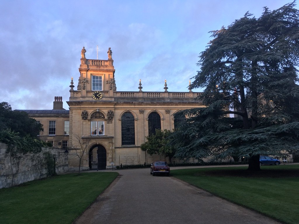 A grand historic building with a clock tower and statues, lit by the evening sun, surrounded by trees and a pathway.