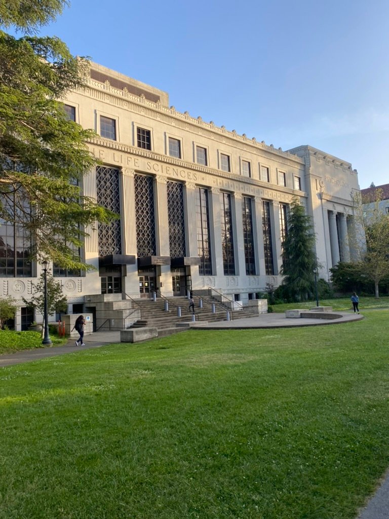 A large academic building labeled “Life Sciences” and “Psychology,” with tall windows and wide front steps, surrounded by grass and a few students walking.